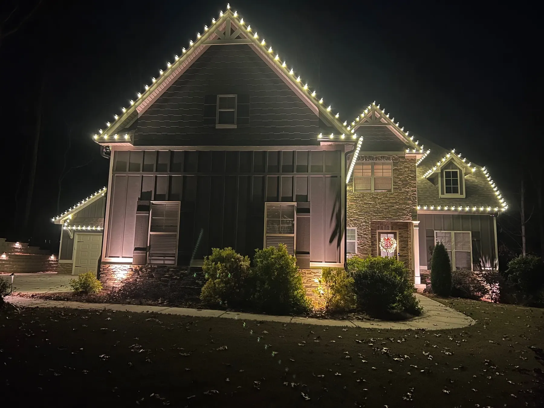 Two-storey home at night with warm white bulbs following the steep roof peaks and dormers, uplighting on the facade and a wreath on the front door